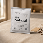 A single foil coffee drips bag labeled 'The Natural' by Brewspecially Coffee, standing upright on a beige fabric surface with a few coffee beans and a small tag nearby. The background is softly blurred and appears to be a warm indoor setting.